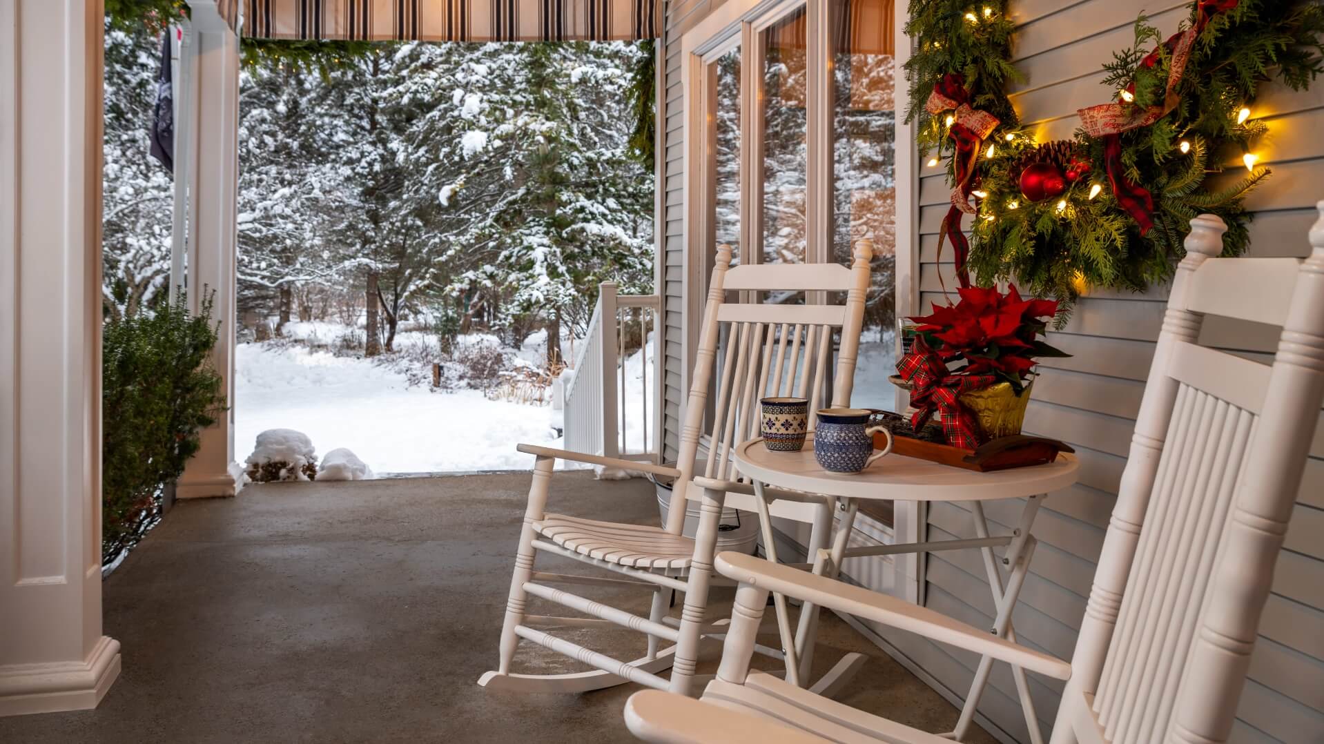 A cozy porch adorned with a festive wreath, rocking chairs, and a snow-covered landscape in the background.