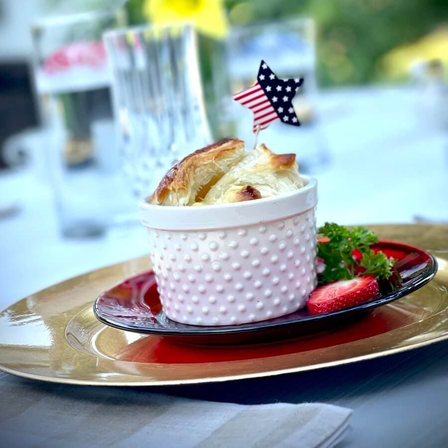 A white ceramic dish with a flaky pastry lid, garnished with a red strawberry and green garnish, topped with a small American flag decoration.